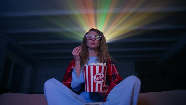 Young woman wrapped in a blanket is enjoys a movie with popcorn. Excited woman watching 3D movie in her home theater, colorful projector beams in the background. High quality 4k footage