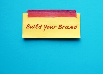 note stick on blue copy space background with handwritten text BUILD YOUR BRAND, means entrepreneur differentiate service product from competitor by creating brand to attract customers perception