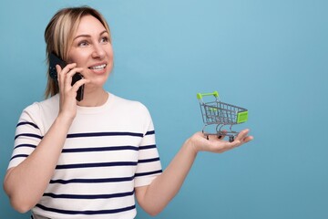 horizontal photo for banner of attractive shopper girl talking on phone holding shopping cart on blue background with copy space