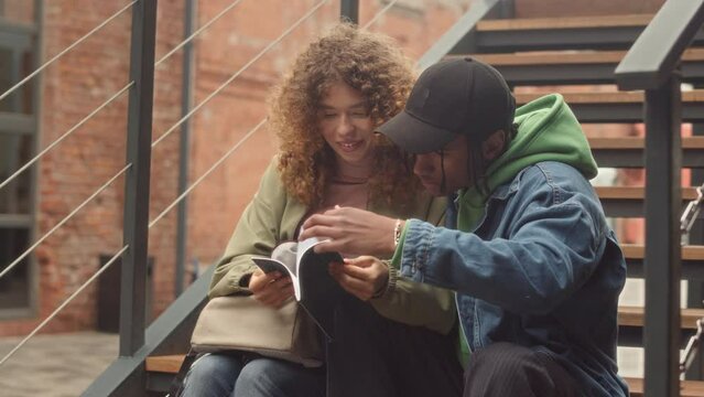 Slowmo of multiethnic romantic couple of college students sitting on stairs outdoors discussing home assignment