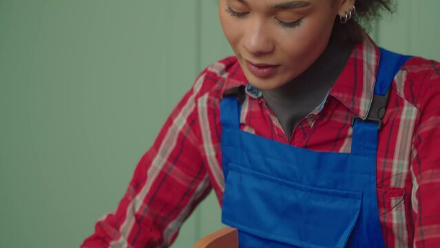 Portrait of skillful busy charming African American young handywoman in protective wear doing DIY, putting together self-assembly wooden furniture using tool in domestic room.
