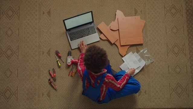 Top view of attractive black handywoman in protective workwear with work and power tools sitting on floor, watching video tutorial online on laptop , learning to assemble DIY furniture at home.