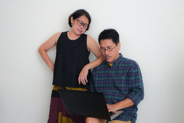 A man working on his laptop, his wife standing nearby, her elbow on her husband's shoulder, both...