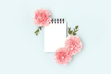 Blank notepad mockup, pink carnation flowers and green eucalyptus branches on a blue background. Place for text.