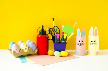 Spring Easter holiday.  Craft, children's creativity, on a colored background on the table are crafts made of colored paper, cute rabbits and Easter eggs, pencils, scissors.  Front view.
