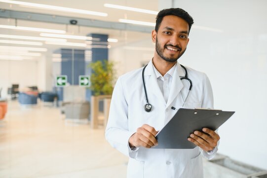 Portrait of male indian doctor wearing white coat having open door on clinic corridor as background
