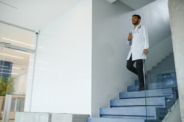 Fototapeta premium Portrait of happy friendly male Indian latin doctor medical worker wearing white coat with stethoscope around neck standing in modern private clinic looking at camera. Medical healthcare concept