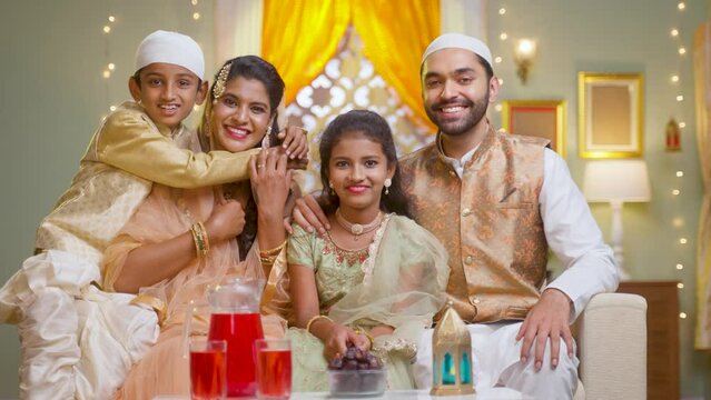 Happy smiling muslim couple with kids in traditional dress by looking at camera during ramdan fest celebration while sitting on sofa at home.