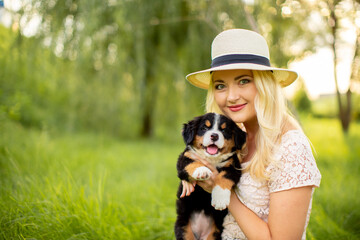 Naklejka na ściany i meble A beautiful girl with a dog sits in the park on the grass. Love to the animals. Home pet on a walk. Animal protection. Bernese mountain dog puppy.