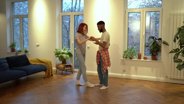 Bonding with loved ones. Happy romantic multiracial young couple in love holding hands enjoying dancing together, dating at home. Smiling joyful multiracial family man and woman learning to dance