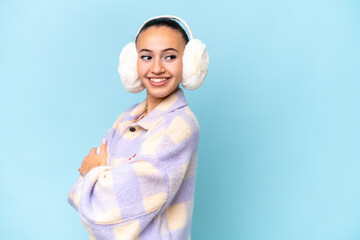 Young Arab woman wearing winter muffs isolated on blue background with arms crossed and happy