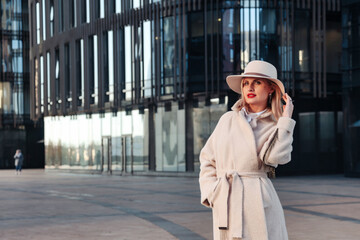 Young lady stylish businesswoman in white coat and hat standing in business district, looking at camera. Fashionable smiling woman near office house at business building background. Copy ad text space