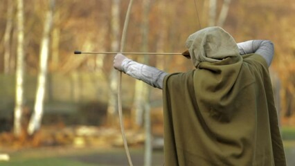 Young man wearing a cloak with bow and arrow in his hands in a forest
