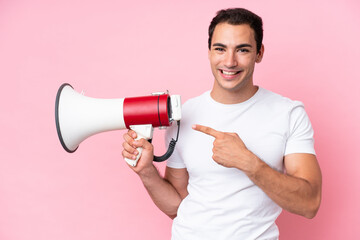 Young caucasian man isolated on pink background holding a megaphone and pointing side