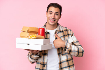 Young caucasian man holding fast food isolated on pink background giving a thumbs up gesture