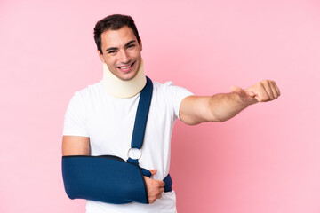 Young caucasian man with broken arm and wearing a sling isolated on pink background giving a thumbs up gesture