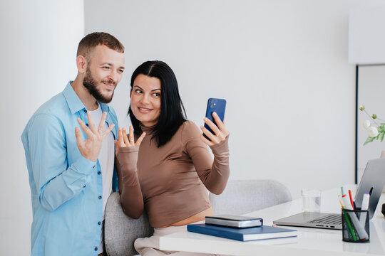 Newlyweds in casual using phone for video call showing wedding rings looking at screen. Happy young caucasian couple at home sharing great news  with parents. Engage, romance. Family.