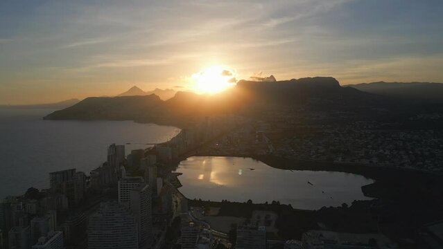 Golden hour over Calpe.Aerial drone view of sun rays,sea,buildings,and reflection in water.Costa Blanca at sunset.Mediterranean sea and Las Salinas lake,mountains at the background.Spain