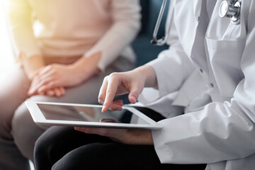 Doctor and patient sitting at sofa in clinic office. The focus is on female physician's hands using tablet computer, close up. Perfect medical service and medicine concept
