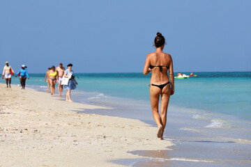 Tanned girl in black bikini walking by a sand on blue sea water and people background. Beach vacation on Caribbean islands