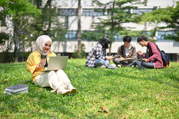 Happy Asian Muslim female college student sitting on the grass, sipping coffee and using her laptop