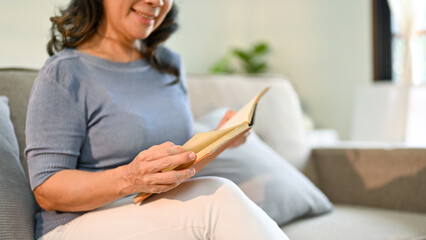 Close-up image an Asian-aged woman reading a book on sofa in her living room.