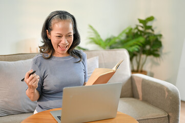Naklejka premium Excited Asian-aged retired woman holding pen and book, using her laptop in living room