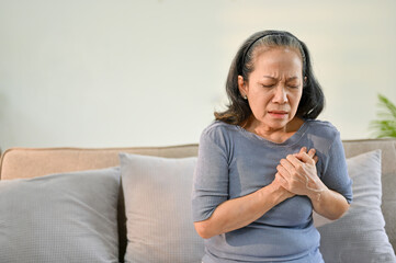 Unwell and sick elderly Asian woman sits on sofa suffering from heart attack or chest pain. - obrazy, fototapety, plakaty