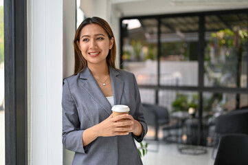 Beautiful Asian businesswoman stands by the window, holds a takeaway coffee cup and daydreams.