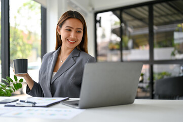 Attractive Asian businesswoman sipping coffee and working on her business tasks on laptop.