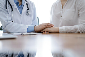 Doctor and patient sitting near each other at the wooden table in clinic. Female physician's hands reassuring woman. Medicine concept