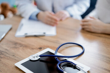 Stethoscope and tablet computer are lying on the wooden table while doctor and patient discussing something at the background. Medicine concept