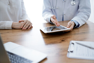 Doctor and patient sitting at the wooden desk in clinic. Female physician's hands pointing into laptop computer monitor, close up. Medicine concept