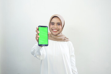 Excited beautiful Asian muslim woman showing green screen mobile phone isolated over white background.