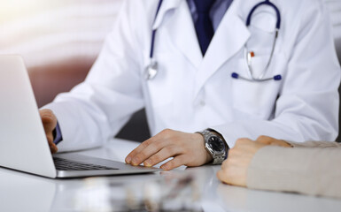 Unknown male doctor and patient woman discussing something while using laptop in a darkened clinic, glare of light on the background