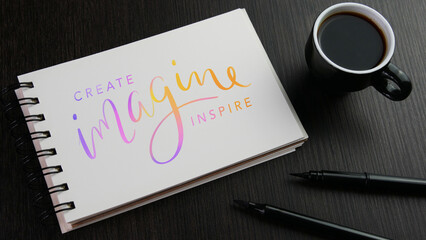 CREATE IMAGINE INSPIRE colorful hand lettering in notebook with cup of espresso and pens on black wooden desk