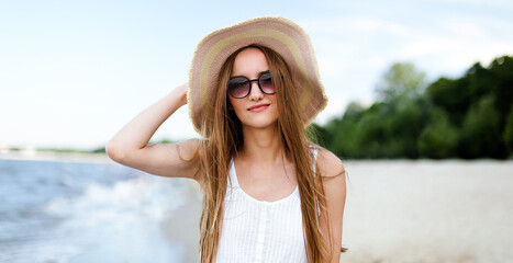 Happy smiling woman in free happiness bliss on ocean beach standing and posing with hat and sunglasses. Portrait of a female model in white summer dress enjoying nature