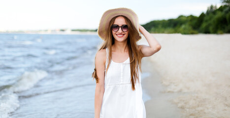 Fototapeta na wymiar Happy smiling woman in free happiness bliss on ocean beach standing and posing with hat and sunglasses. Portrait of a female model in white summer dress enjoying nature