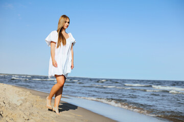 Fototapeta na wymiar Happy smiling beautiful woman is walking on the ocean beach in a white summer dress