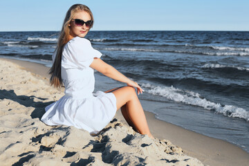 Happy, beautiful blonde woman is sitting on the ocean beach in a white summer dress and sunglasses