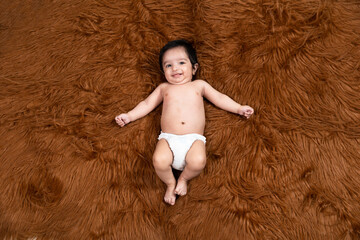Cute smiling little indian baby wearing diaper lying on brown fur background. Asian two months old newborn baby.