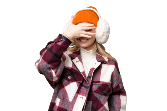 Young  English woman wearing winter muffs over isolated background covering eyes by hands and smiling