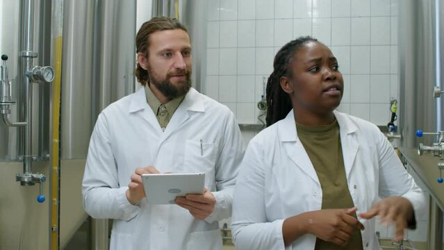 Medium follow shot of Black female and Caucasian male brewery technologists in white coats walking together in production facility, and discussing batches of beer brewing in fermenter tanks