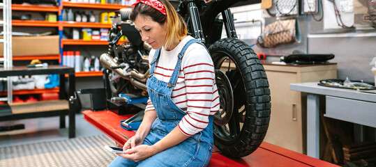 Fototapeta na wymiar Mechanic woman looking phone sitting over platform with motorcycle on factory