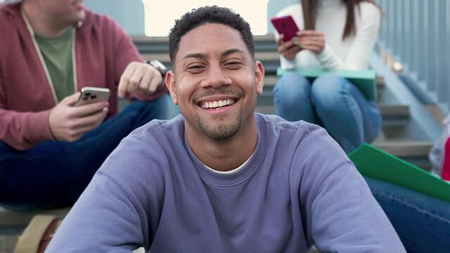 Happy young latino hispanic student man smiling at camera at college campus sitting on staircase with classmate friends on the background using mobile phone and social gathering