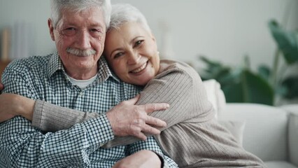 Senior couple relaxing hugging sitting on couch in living room at home, elderly wife and husband in lifelong relationships, happy marriage concept. Old gray haired people smile and look at camera.