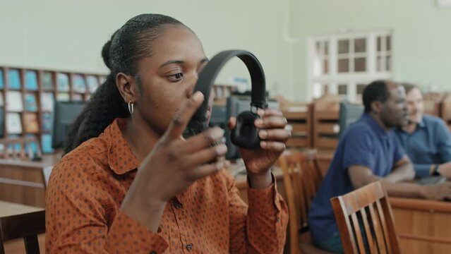 Medium close-up selective focus shot of young Black female student working ion computer in college library putting on headphones