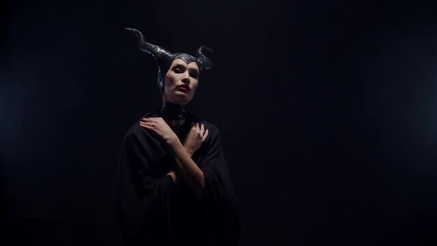 An evil witch villain woman from a fairy tale with horns stands with her arms crossed on her chest, turns and looks at the camera in a dark black studio. Cartoon, fantasy film