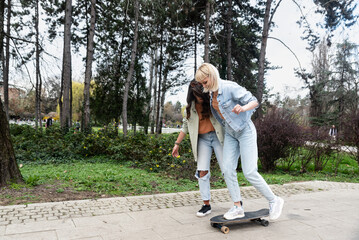 Fototapeta premium Two cool-looking hipster friends in park learning to ride a skateboard, having fun between studying or working, spending time together in a nice sunny weather. A friend teaches her roommate to skate