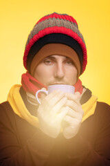 A young man in warm clothes, several hats on a yellow background holds a cup of tea warms himself during a cold
